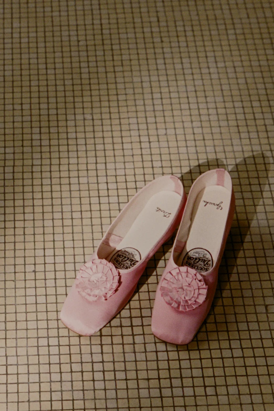 Pair of pink soulier shoes with floral rosette decoration on a warm checkered tile floor.