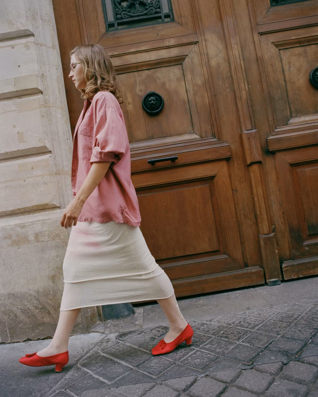 A woman walking past large wooden doors on a cobblestone street, wearing a pink short-sleeve blouse, a cream midi skirt, and red low-heeled pumps. Souliers Dulion, low heeled 1800s souliers designed in Paris and handcrafted in Italy