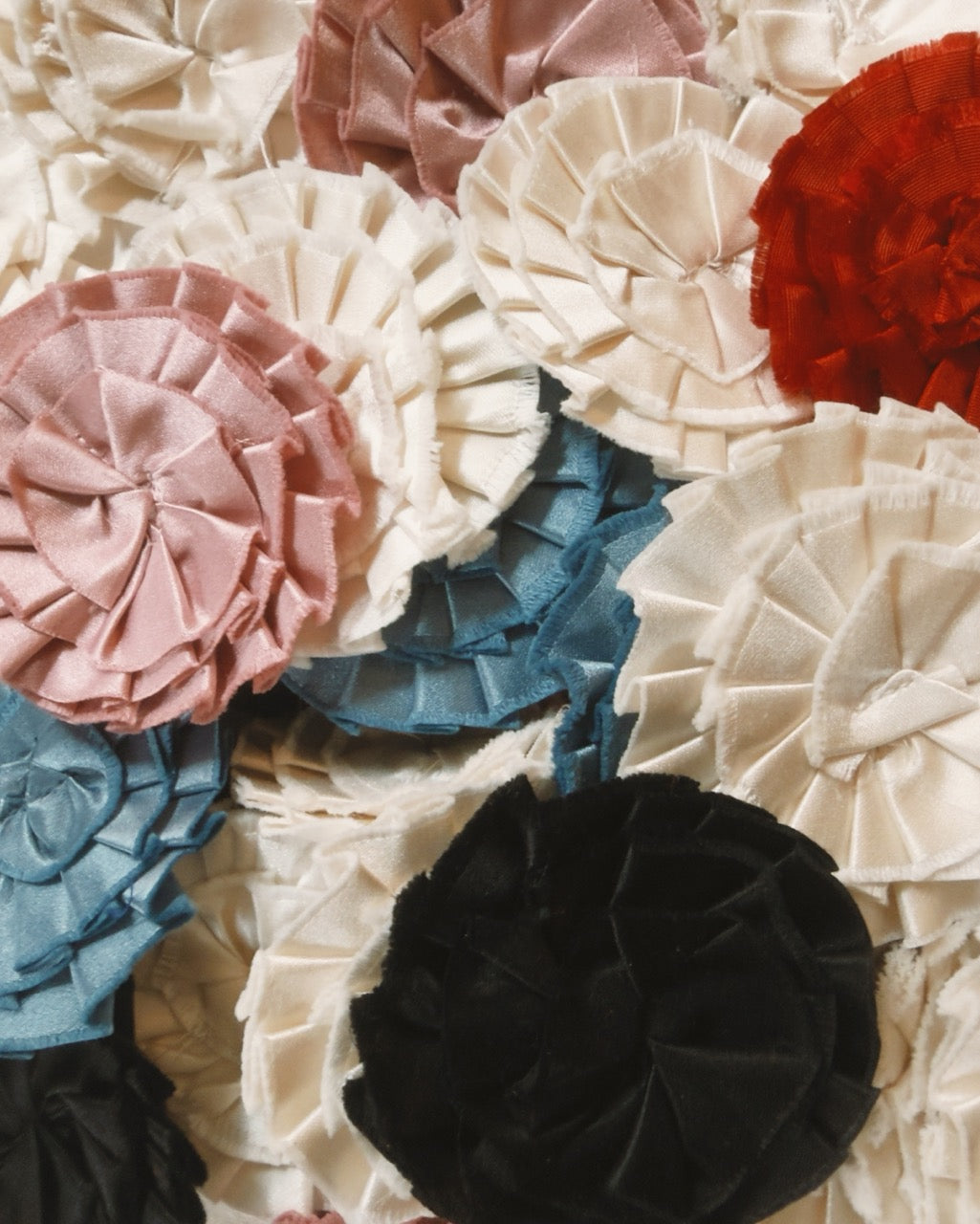 A close-up arrangement of layered ruffled fabric rosettes in cream, blush pink, dusty blue, rust red, and black.