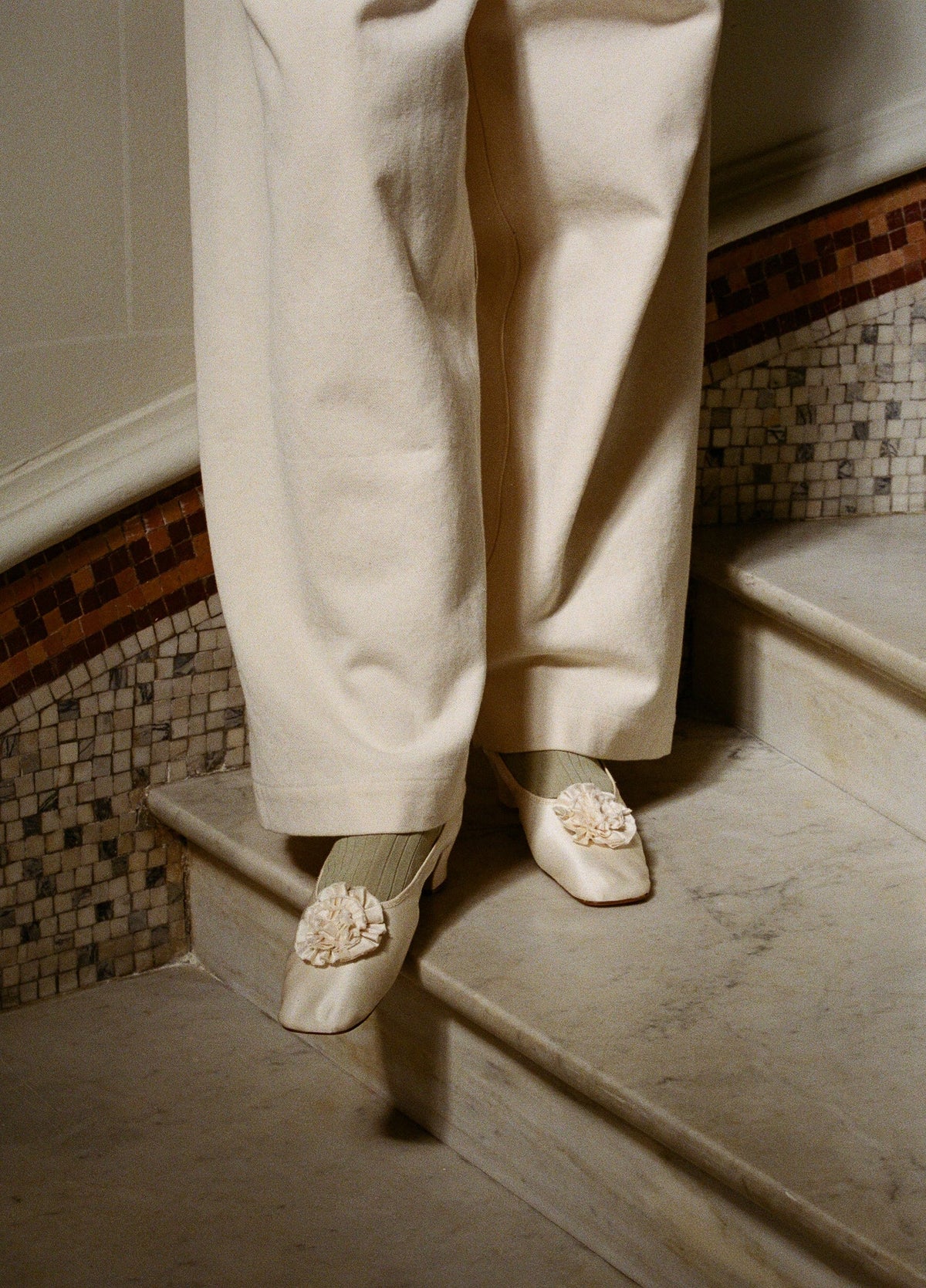A person wearing cream wide-leg trousers and matching cream satin pointed shoes with floral embellishments, standing on stone stairs. Souliers Dulion, low heeled 1800s souliers designed in Paris and handcrafted in Italy